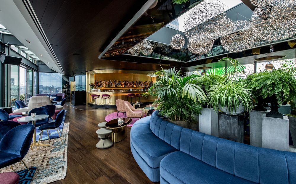 Sea Containers London Hotel Exterior photo The photo depicts a stylish interior of a lounge or bar area. There are modern furnishings, including a large, curved blue sofa, and various seating arrangements such as chairs and tables. The space features lush green plants that add a touch of natu