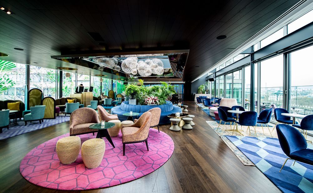 Sea Containers London Hotel Exterior photo The photo shows a modern and stylish interior of a lounge or café space. It features a combination of colorful furniture, including plush chairs and small tables arranged on a vibrant pink rug. There are large windows allowing natural light to flood 